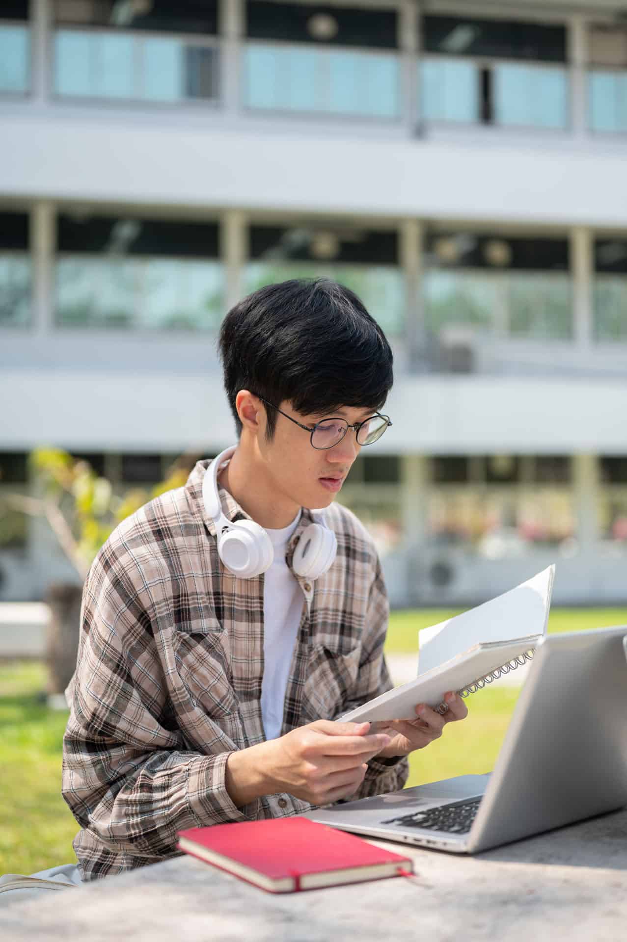 A university-aged student wearing glasses, reading notes outside on a campus bench.