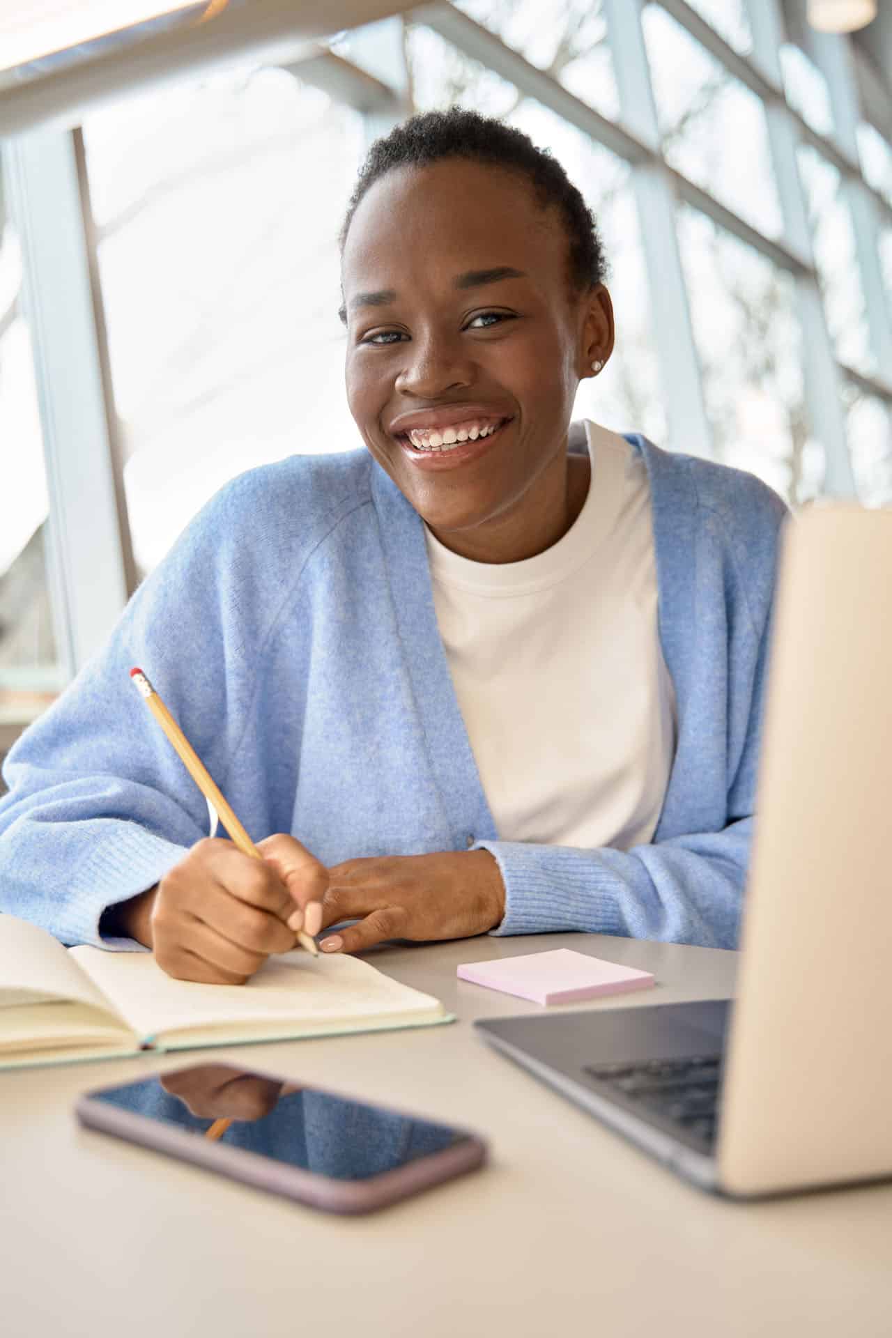 A student smiling at a desk with a laptop and notebook, readying application documents.